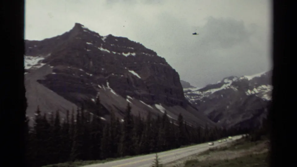 1979 Canada mountain road scene with small cars traveling through a valley beneath steep rocky slopes and evergreen forest.