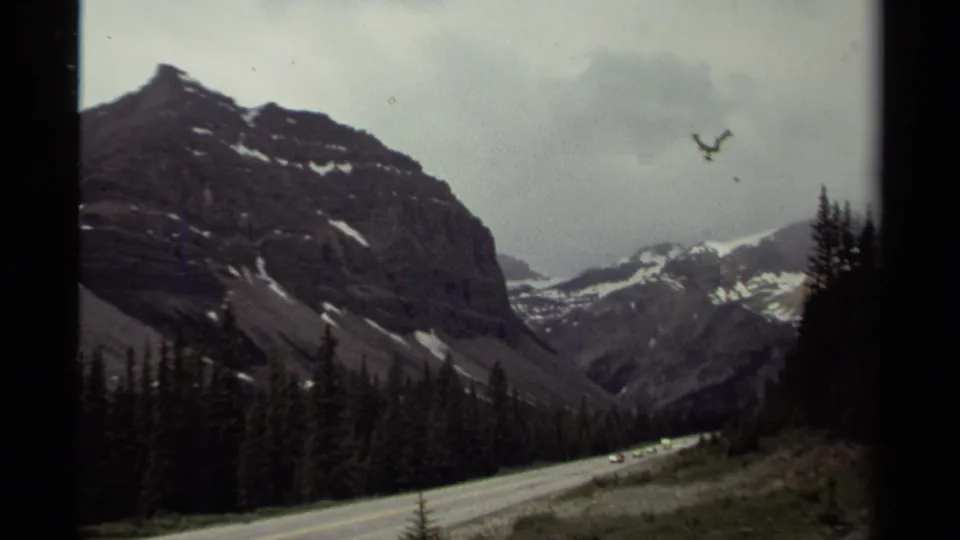 A 1979 Canada home-movie frame shows cars on a winding road through a mountain valley, with dark conifers and distant snow patches.