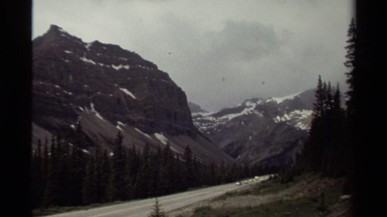1979 Canada 8mm frame showing a paved mountain road, dense conifers and snow-streaked peaks under an overcast sky.