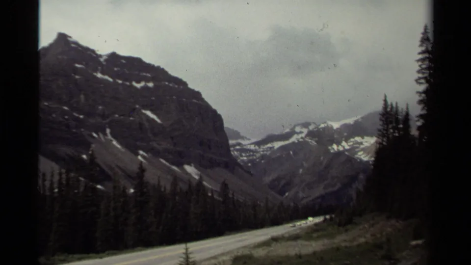 Canadian mountain landscape on 8mm film, with a road edge, dense trees and distant snow patches on high peaks.