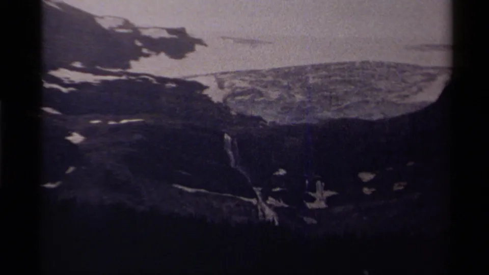 8mm frame from Canada showing a darker mountain view with snowfields, shadowed forest and a small bright streak near the slope.