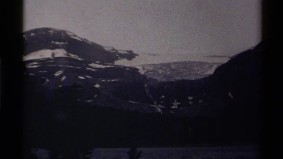 Canadian 8mm frame showing a snow-marked mountain slope, dark valley edge, and faint lake surface in low evening or night light.