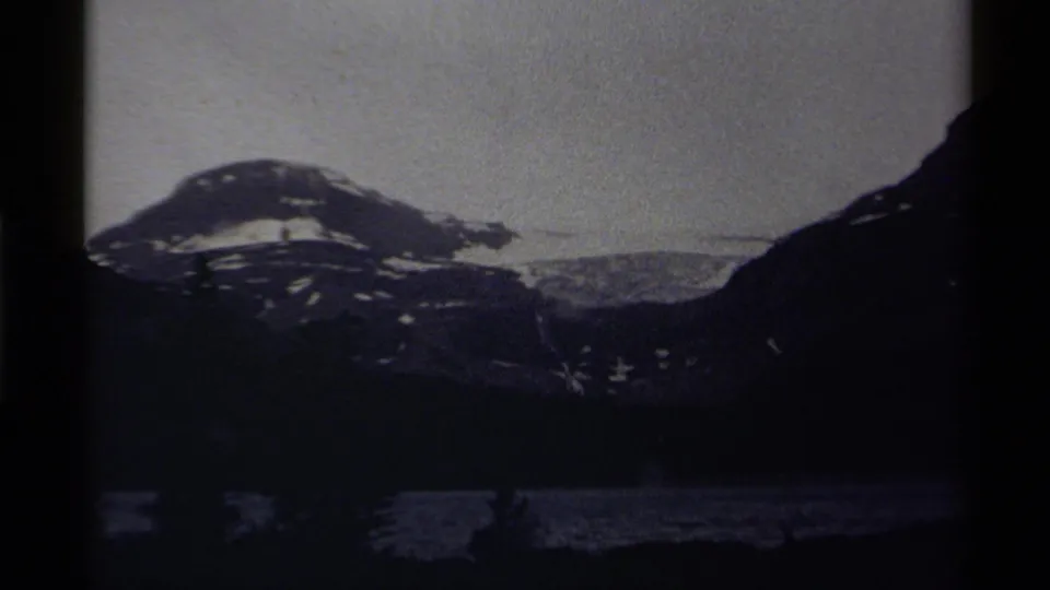 Low-light 8mm frame from Canada showing a snow-dusted mountain mass, shadowed lake, and pale sky near the end of the clip.