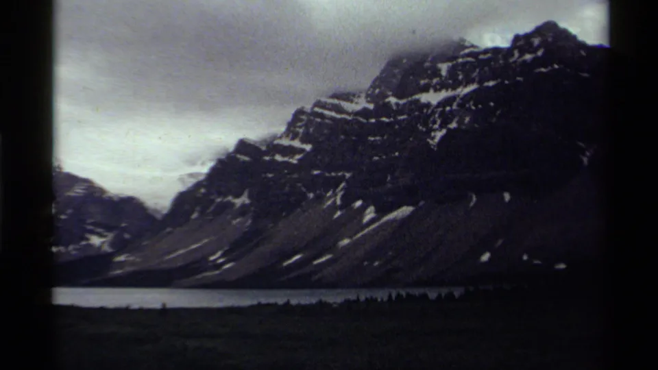 Vintage 8mm Canada frame with a large shadowed mountain, patches of snow, a narrow lake, and muted clouds.