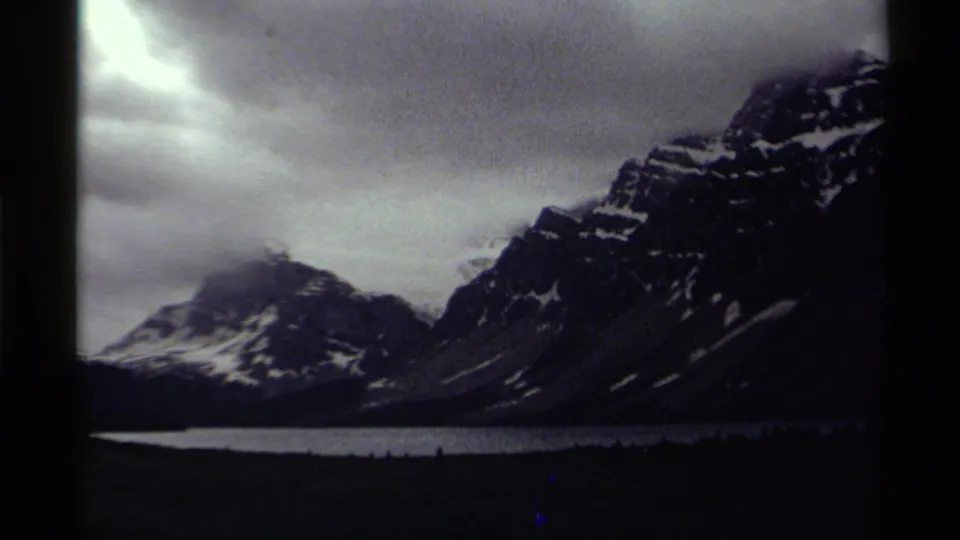 Vintage 8mm Canada frame of a dark mountain ridge, bright lake strip, forested base, and low gray clouds.