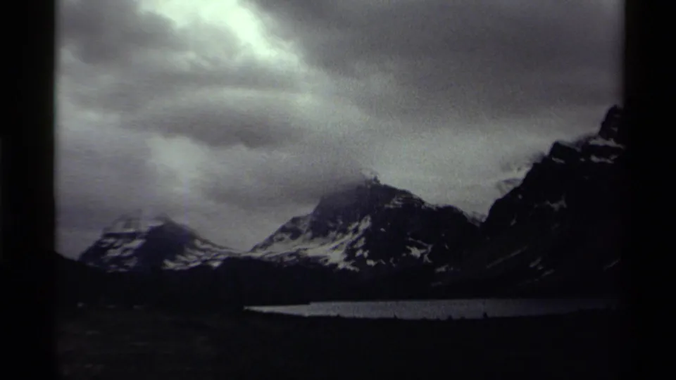 Archival 8mm Canada landscape with snowy mountain slopes, lake water, and shifting light in heavy clouds.