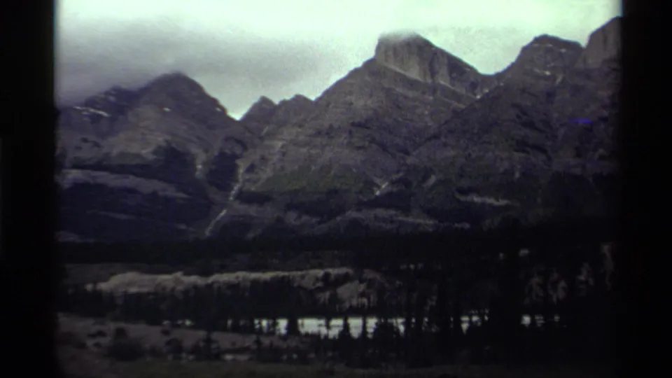1979 Canada 8mm frame showing dark rocky peaks, low cloud, forest, and a narrow reflective body of water in the valley.