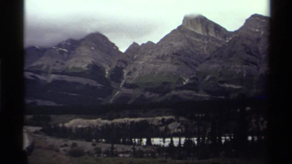 1979 Canada 8mm frame of a broad mountain valley with ridged slopes, evergreen forest, and bright water near the lower edge.