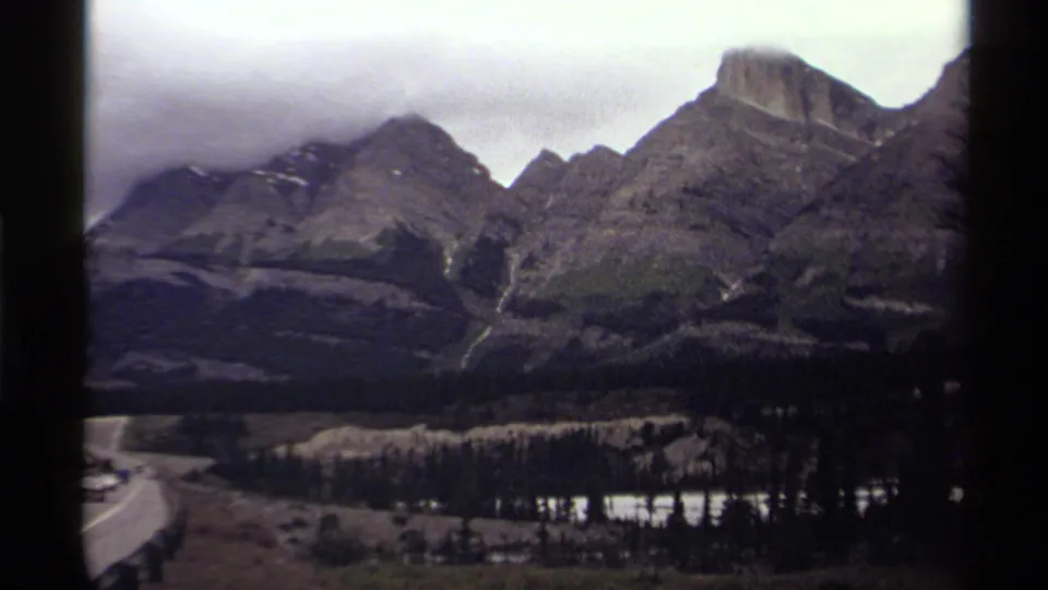 1979 Canada 8mm frame showing cloudy mountain peaks, a forested valley, water, and a winding road at the lower left.
