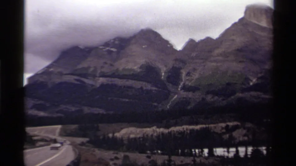 1979 Canada 8mm frame of a winding road curving through a valley beneath mountains, forest, and a small reflective lake or river.