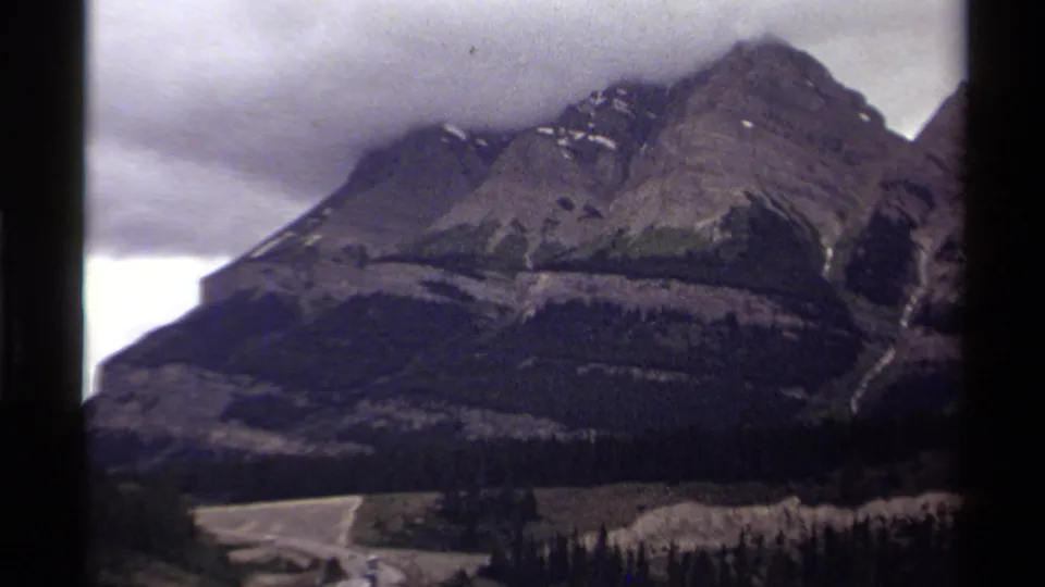 1979 Canada home-movie view of a high rocky peak, low cloud cover, forested slopes, and a road at the base.