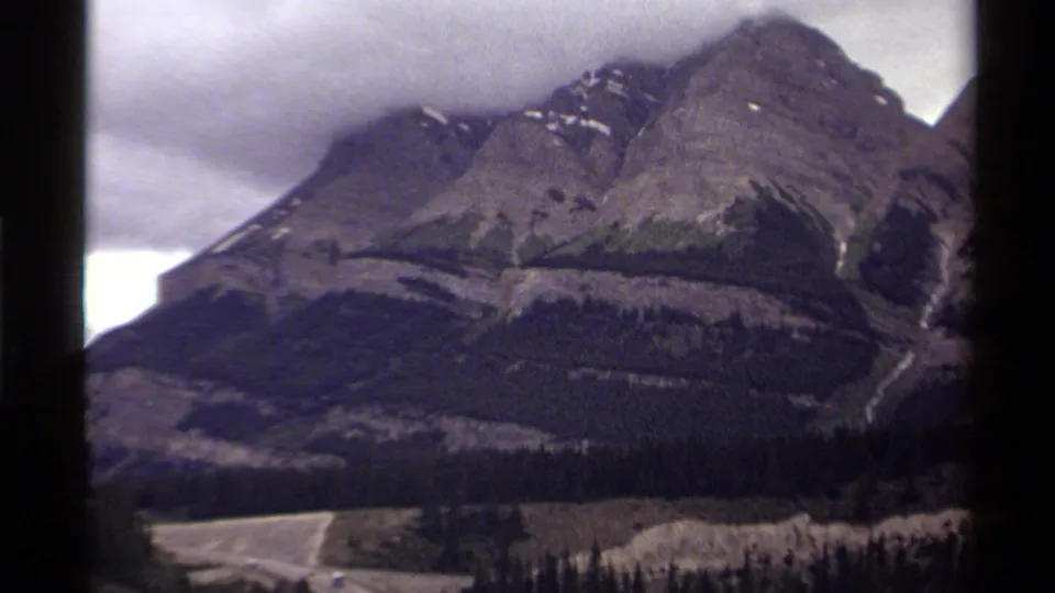 1979 Canada 8mm frame with a closer mountain slope, dark evergreen forest, pale road, and heavy cloud cover.