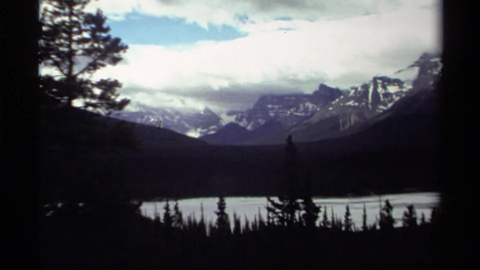 1979 Canada home-movie frame showing a lake, dark tree silhouettes, distant snow-capped mountains, and a partly cloudy sky.