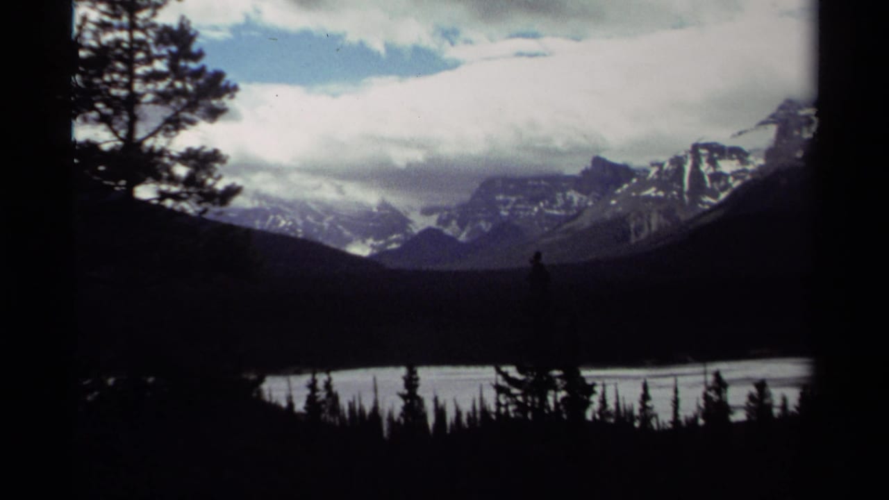 1979 Canada 8mm frame showing snow-streaked mountains beyond lake water, with dark conifers in the foreground and heavy clouds overhead.