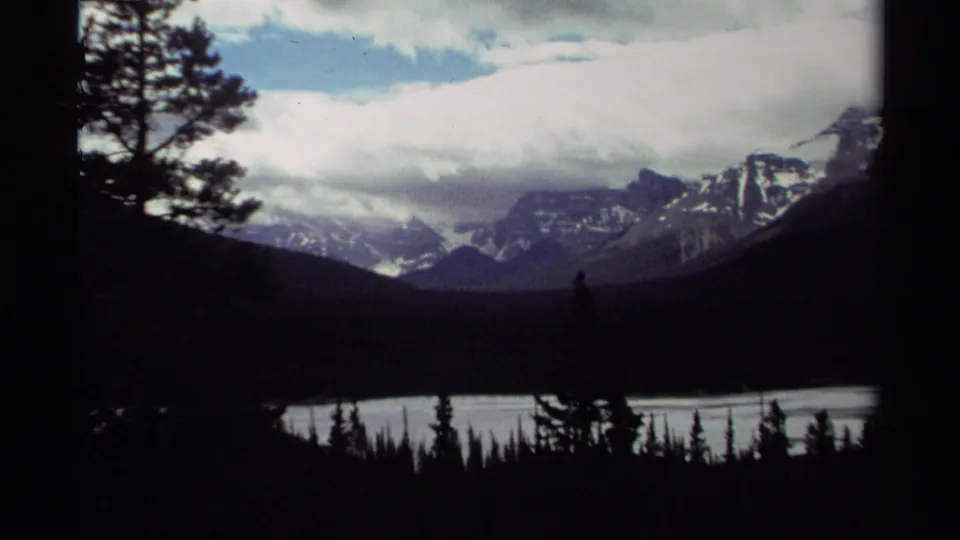 1979 Canada 8mm frame showing snow-streaked mountains beyond lake water, with dark conifers in the foreground and heavy clouds overhead.