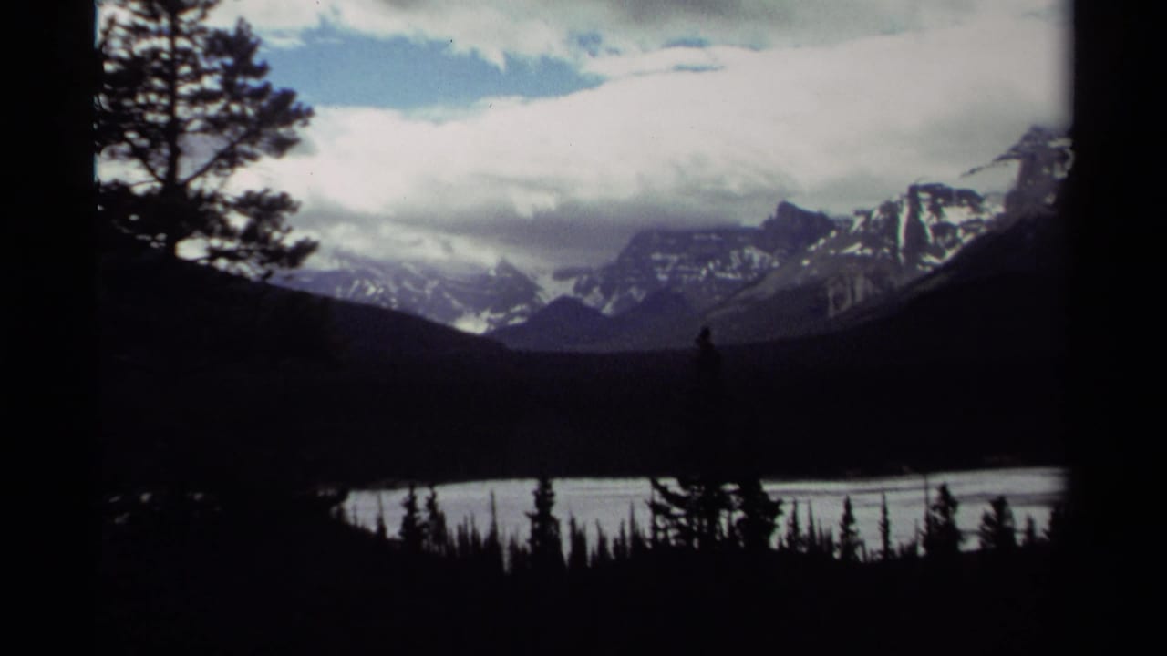 1979 Canada 8mm view of a mountain lake, distant snow-streaked peaks, silhouetted trees, and layered clouds across the sky.