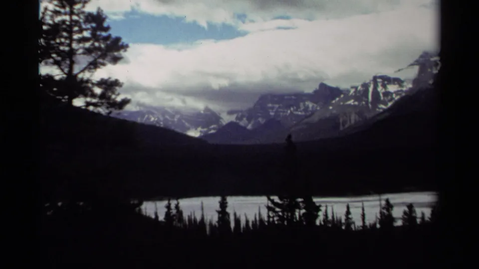 1979 Canada 8mm view of a mountain lake, distant snow-streaked peaks, silhouetted trees, and layered clouds across the sky.