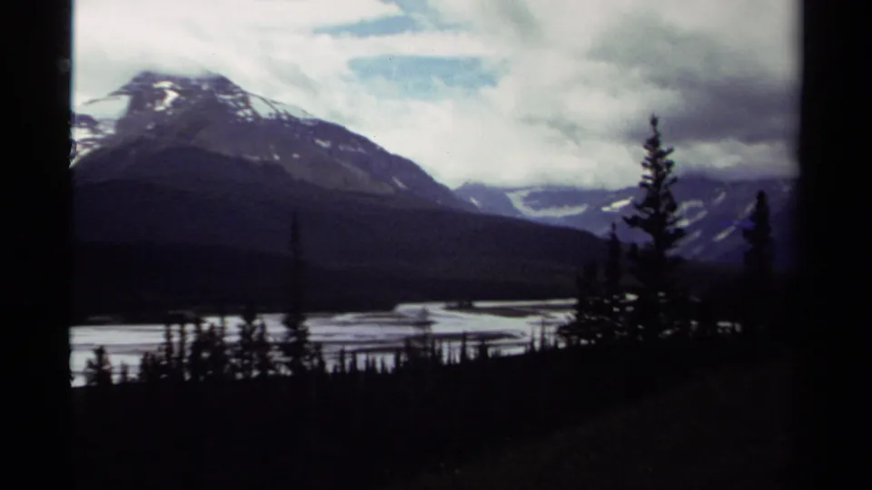 1979 Canada 8mm frame showing a broad mountain and valley waterway, with foreground conifers and low clouds softening the snowy slopes.