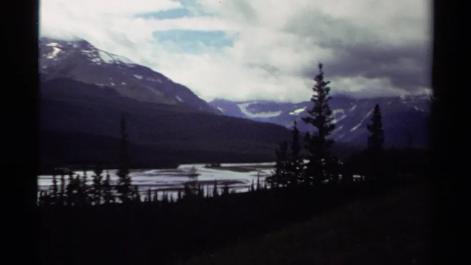 1979 Canada 8mm frame of evergreen silhouettes above a lake or river, with layered mountains and a bright break in the cloudy sky.