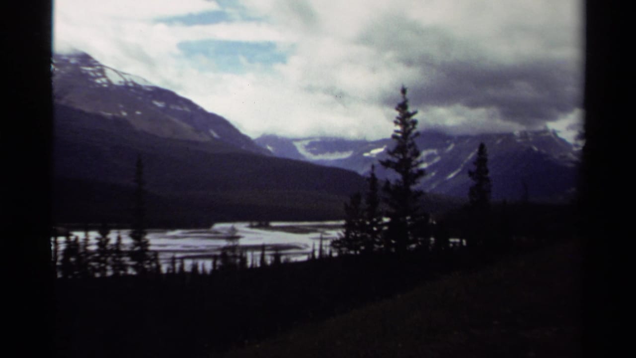 1979 Canadian 8mm frame showing snow-streaked mountains, water, and dark evergreens beneath heavy clouds.