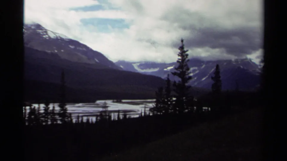1979 Canadian 8mm frame showing snow-streaked mountains, water, and dark evergreens beneath heavy clouds.