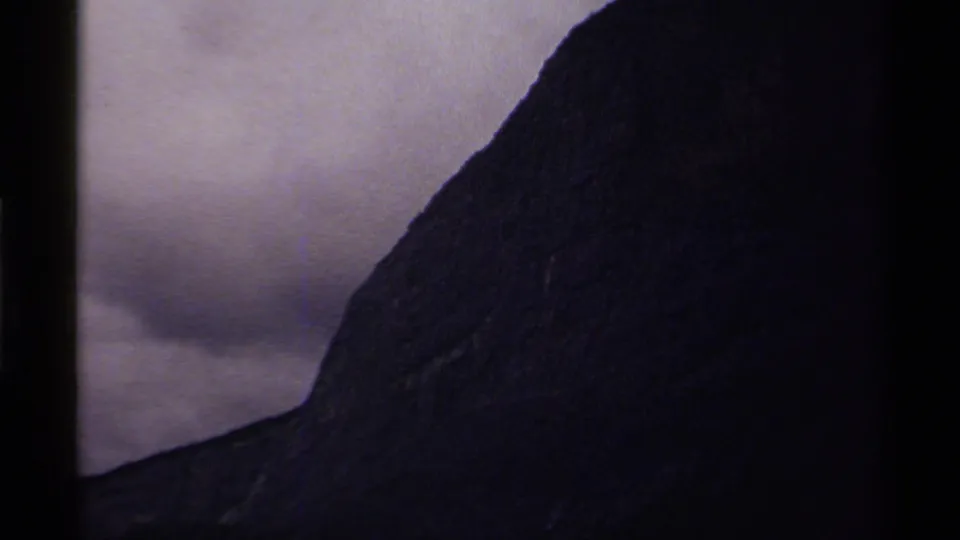 8mm frame of a Canadian mountain slope, mostly in shadow, with gray cloud cover behind it.