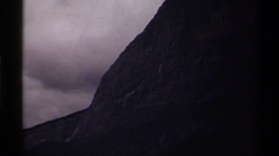 Late-1970s Canadian home-movie frame showing a dark mountain wall under a muted cloudy sky.