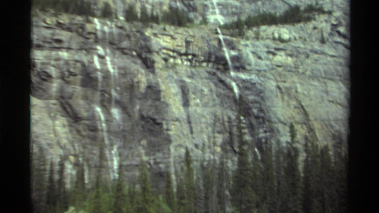 1979 Canada 8mm frame showing a rocky mountain face with narrow waterfalls and dark evergreen trees on ledges.