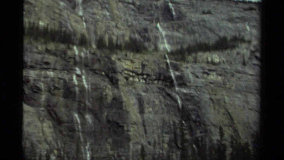 1979 Canada home-movie frame of a distant rocky cliff with several pale waterfalls and evergreen trees along the ridge.