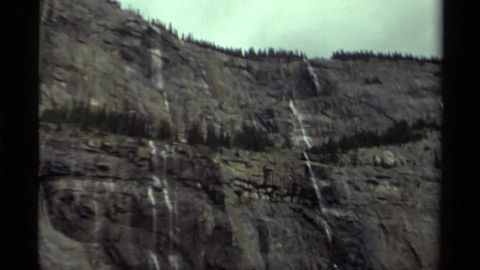 1979 Canada 8mm frame showing a broader mountain slope with cliff ledges, waterfalls, evergreens, and pale cloudy sky.