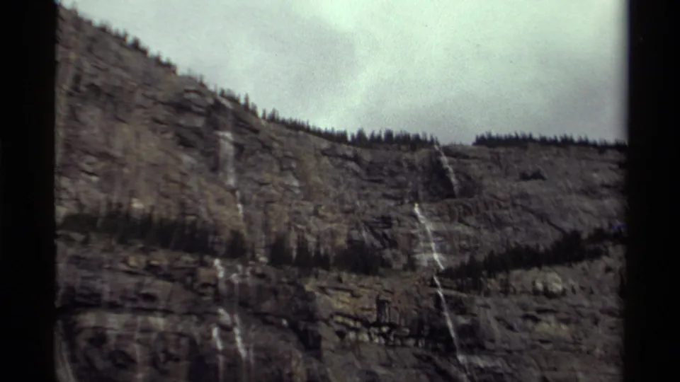 1979 Canada home-movie frame of a rocky mountain wall with thin waterfalls, ridge trees, and an overcast sky.