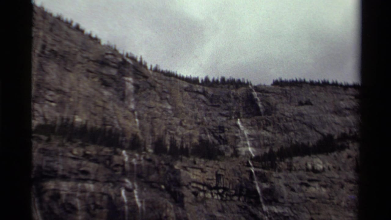1979 Canadian 8mm frame showing a rocky mountain face with narrow waterfalls, scattered evergreens, cloudy sky, and visible film borders.