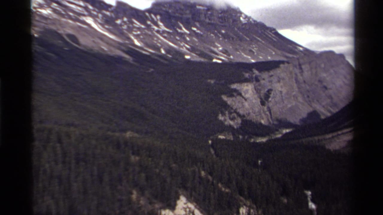 Snow-streaked mountain ridge above a forested valley and pale cliff under cloudy sky.