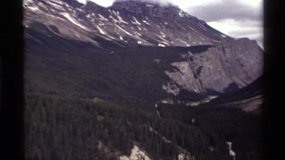1979 Canadian 8mm view of snow-streaked peaks, a dense evergreen valley, and a light rocky cliff beneath broken cloud cover.