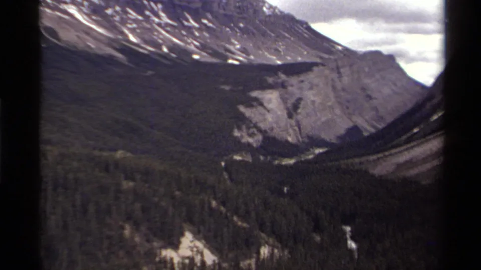 1979 Canadian 8mm frame looking across an evergreen valley toward snow-patched mountains and pale cliffs, with soft grain and film borders.