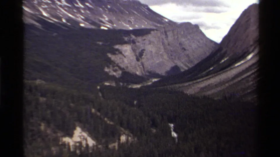 1979 Canadian 8mm view of a dark evergreen valley, pale cliff wall, and snow-marked mountain slopes framed by visible film borders.