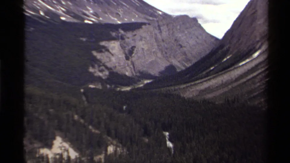 1979 Canada home-movie frame with a closer look at trees, a winding pale route or stream, and steep mountain walls.