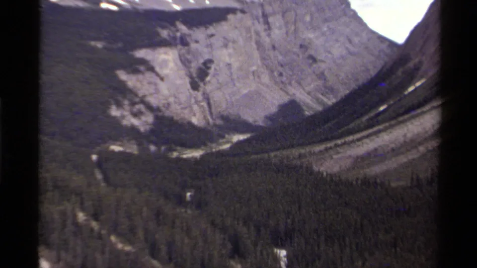 1979 8mm Canada footage showing evergreen forest across a valley beneath steep rocky mountain faces and small snow patches.