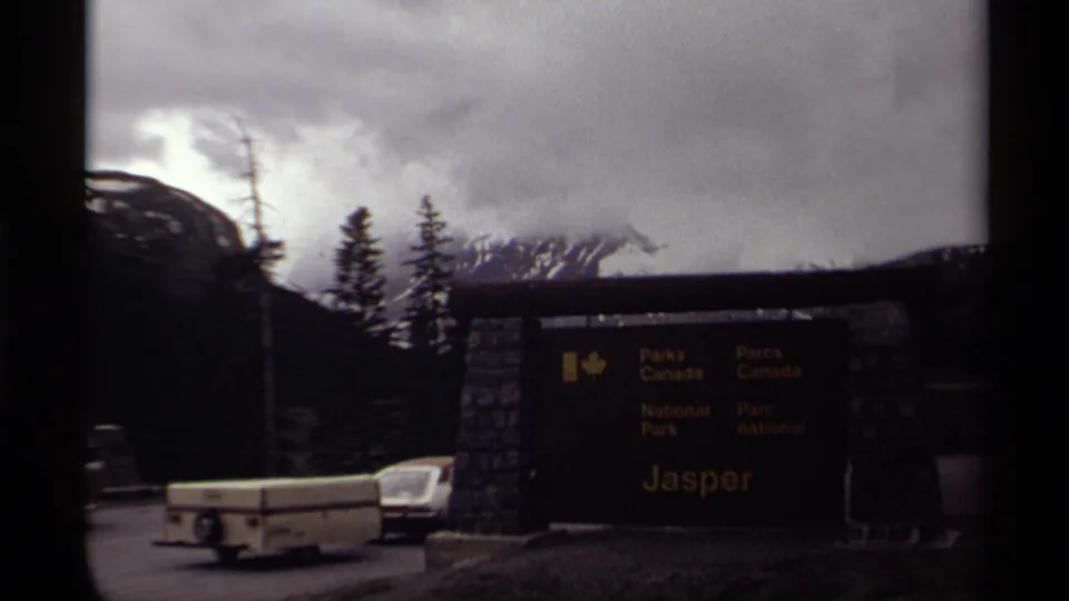 Parks Canada Jasper National Park entrance sign appears beside a road with a camper trailer and car.