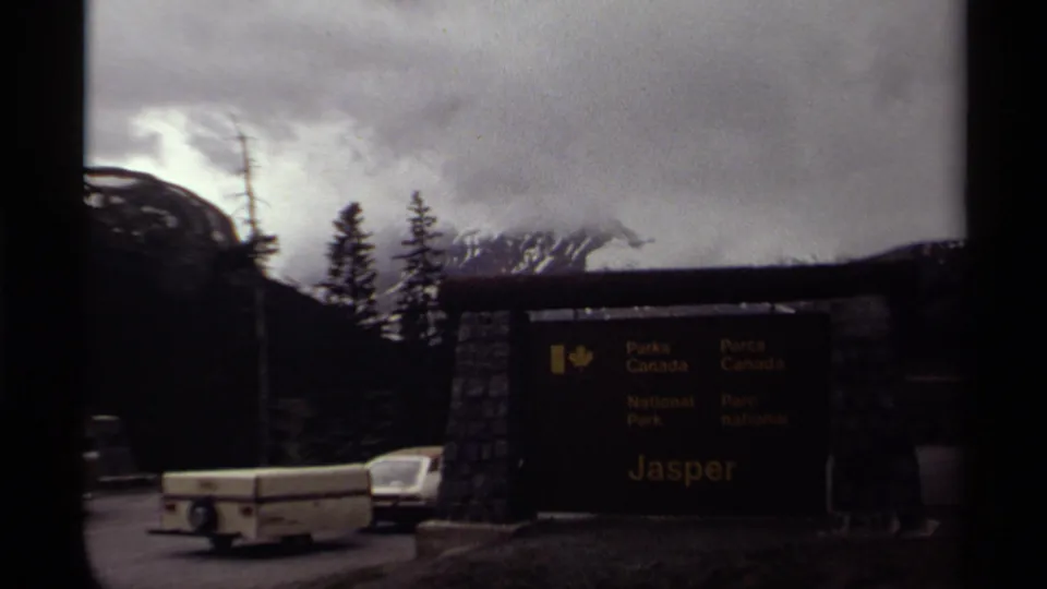 The Jasper National Park entrance sign stands near a road, with mountains, trees, and a cloudy sky behind it.