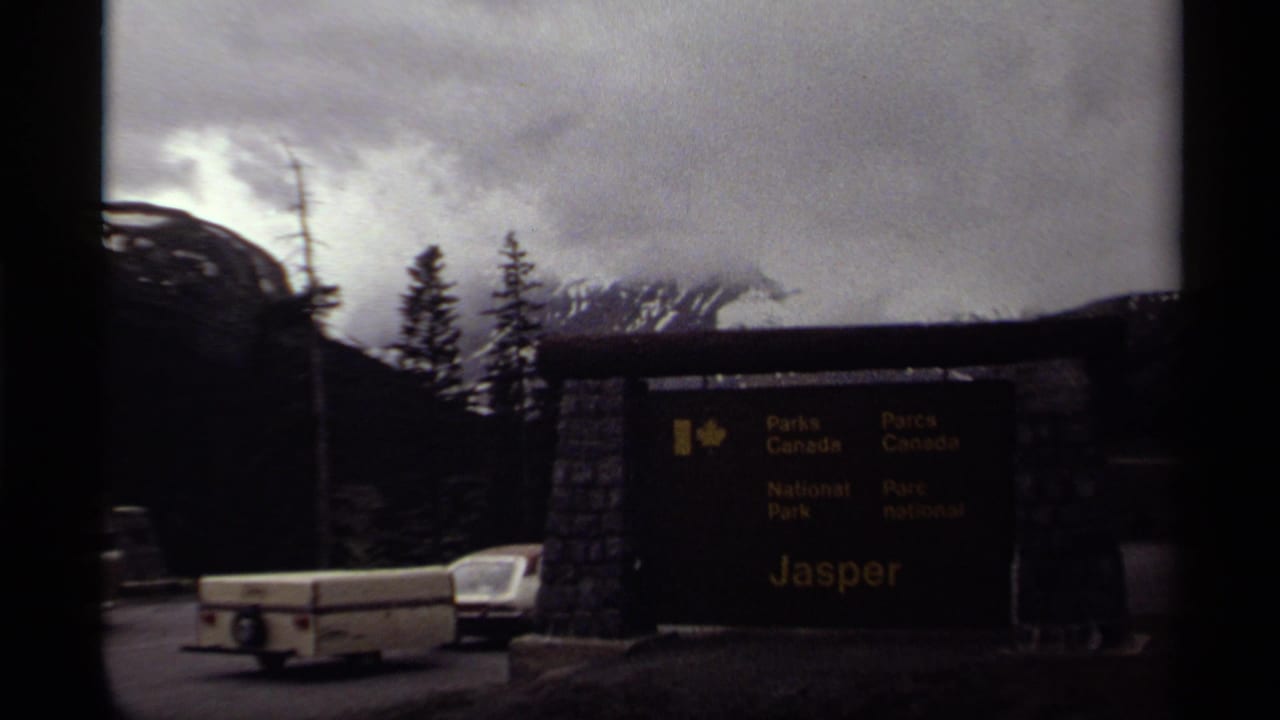 1979 8mm frame of the Jasper National Park entrance sign in Canada, with a vintage car and trailer near evergreen trees and cloudy mountains.
