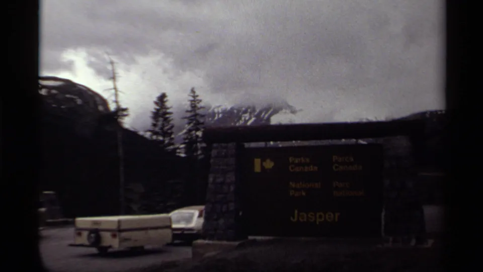 1979 8mm frame of the Jasper National Park entrance sign in Canada, with a vintage car and trailer near evergreen trees and cloudy mountains.