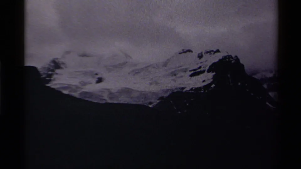 Grainy 8mm view of a Canadian Rockies peak, with snowfields above a dark foreground ridge and heavy cloud cover.