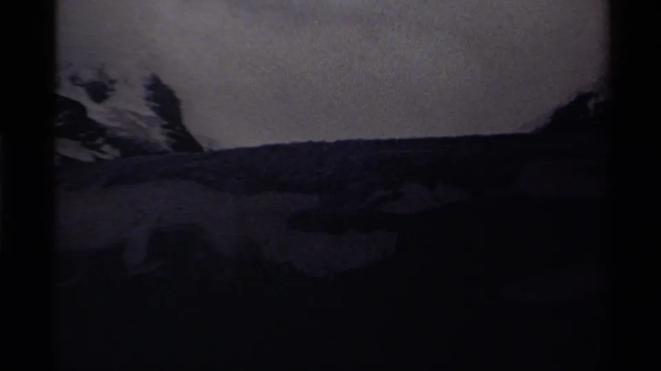 1979 Canadian 8mm frame looking across dark foreground ice or water toward a snow-covered mountain side and muted sky.