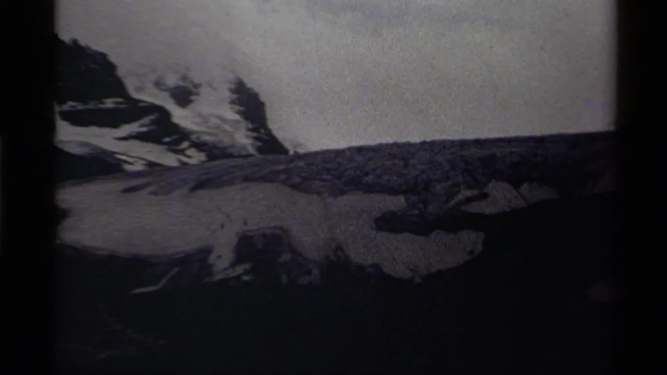 1979 Canadian 8mm frame showing dark rock faces, snowfields, and a distant mountain ridge under cloud cover.