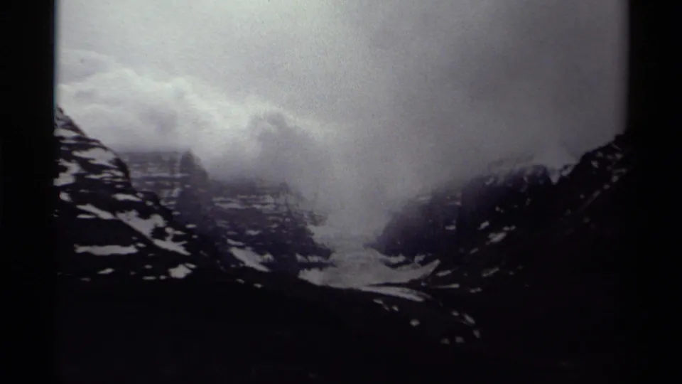 Canadian mountain landscape on 8mm film, with low cloud cover, dark ridgelines, and scattered snow patches.