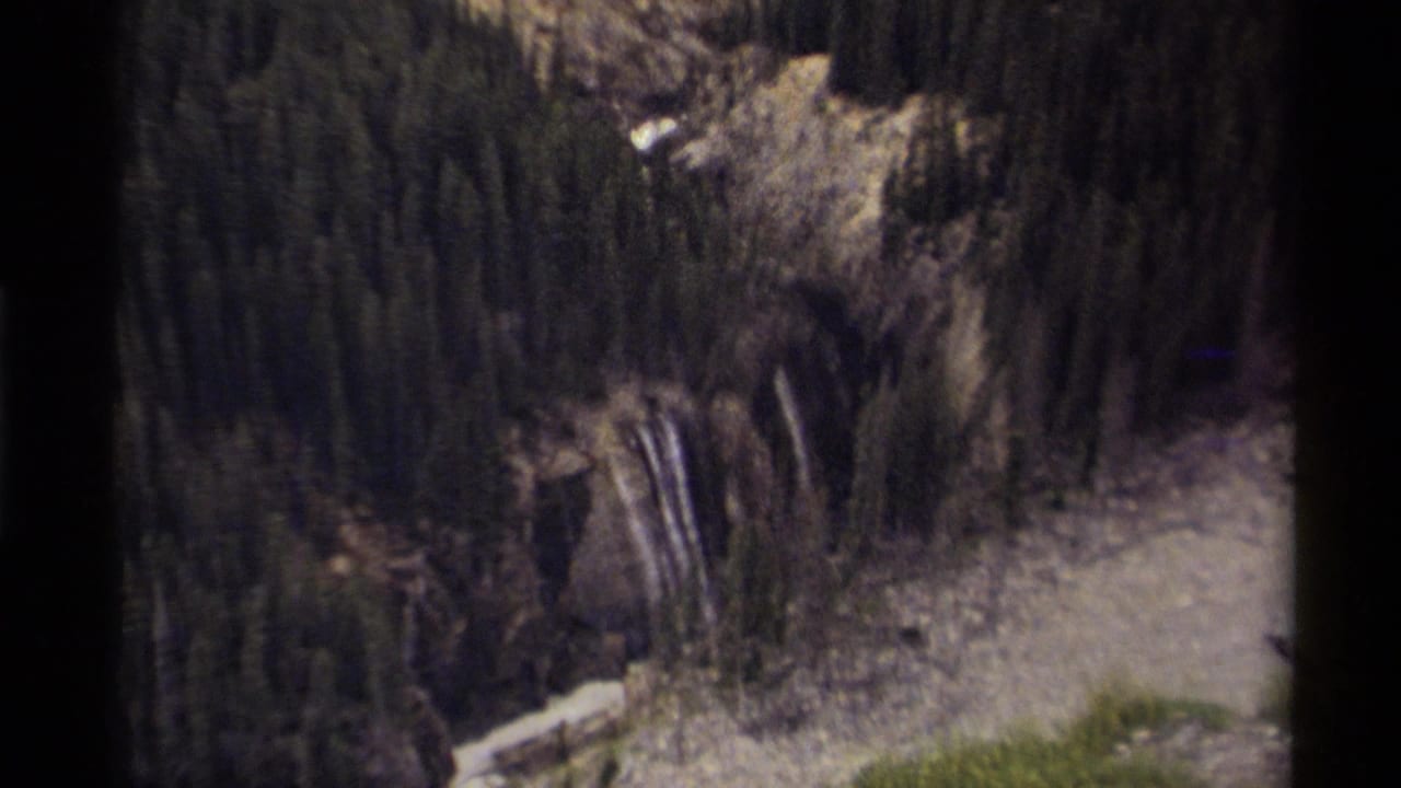 1979 Canadian 8mm aerial view looking down over evergreen forest, rocky slopes, a waterfall, and a pale river channel.