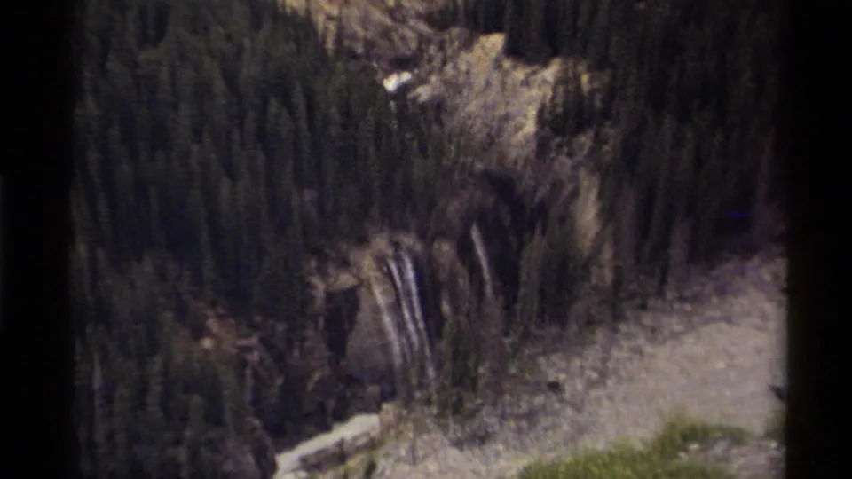 1979 Canadian 8mm aerial view looking down over evergreen forest, rocky slopes, a waterfall, and a pale river channel.