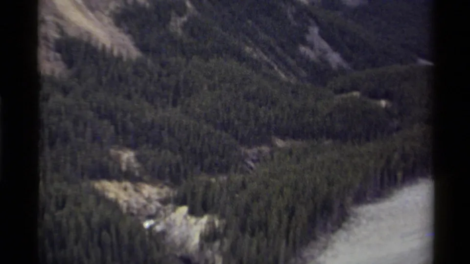 1979 Canadian 8mm aerial frame with evergreen forest, pale river water, rocky slopes, and a partial waterfall area at the edge of view.