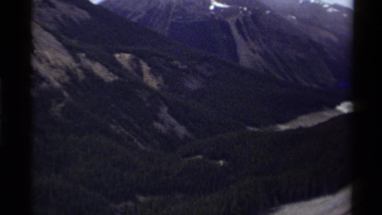 1979 Canada 8mm frame showing a shadowed mountain valley, snow-streaked ridges, forest, and pale water below.